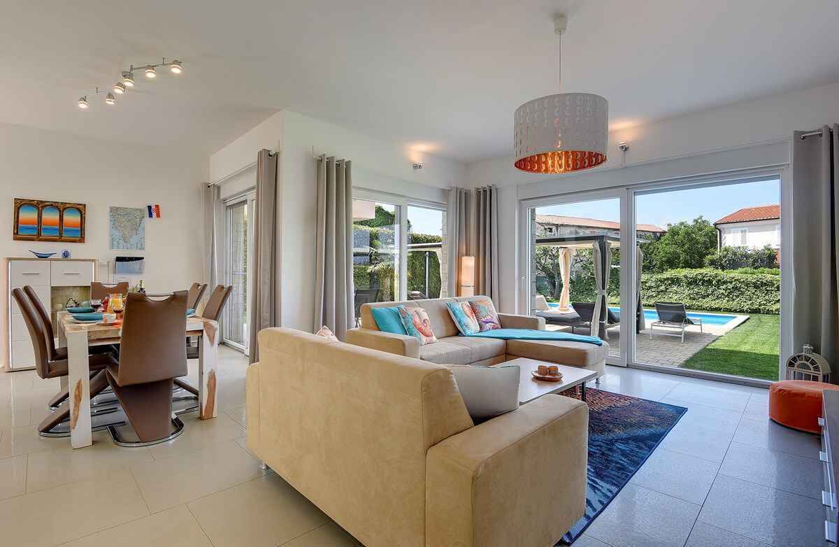 A modern living room with a sofa, dining table, and chairs. There is a large window with a view of a garden and a pool.