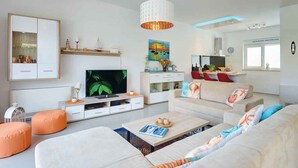 A modern living room with a flat-screen TV, white sofas, orange ottomans, and a large painting.