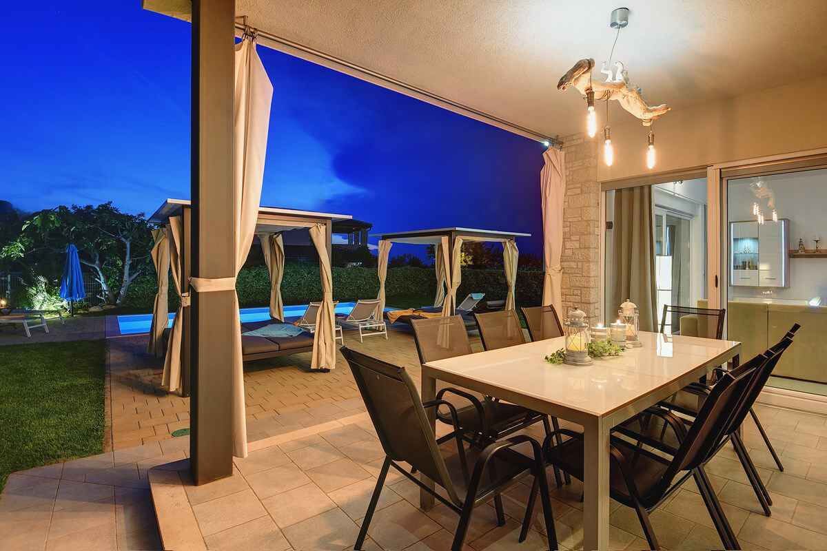 Uma área de refeições ao ar livre com mesa e cadeiras, piscina e um espaço de estar coberto com guarda-sóis.