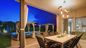 An outdoor dining area with a table and chairs, a pool, and a covered seating area with umbrellas.