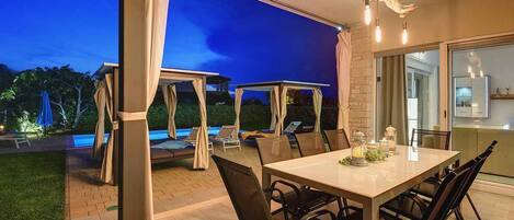 Uma área de refeições ao ar livre com mesa e cadeiras, piscina e um espaço de estar coberto com guarda-sóis.