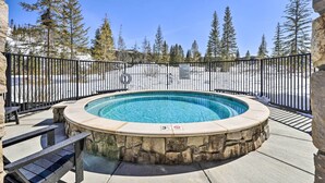 A hot tub surrounded by stone walls and a black metal fence, with a clear view of snow-covered ground and tall evergreen trees in the background. - Modern Winter Park Condo w/ Community Hot Tub! (Winter Park)