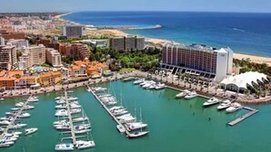 A marina with numerous boats docked, a large hotel building, and a beach in the background. - Modern 2 bedroom Vilamoura Marin (Quarteira)