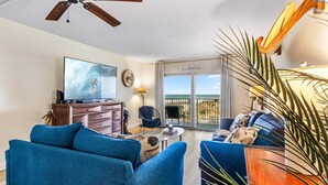 Een woonkamer met een televisie, blauwe banken, een plafondventilator, een balkon met uitzicht en een bordje met de tekst 'Our Happy Place'.