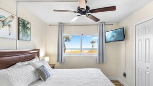 Een slaapkamer met een bed, een plafondventilator, een televisie, een raam met uitzicht op het strand en kunst aan de muur.