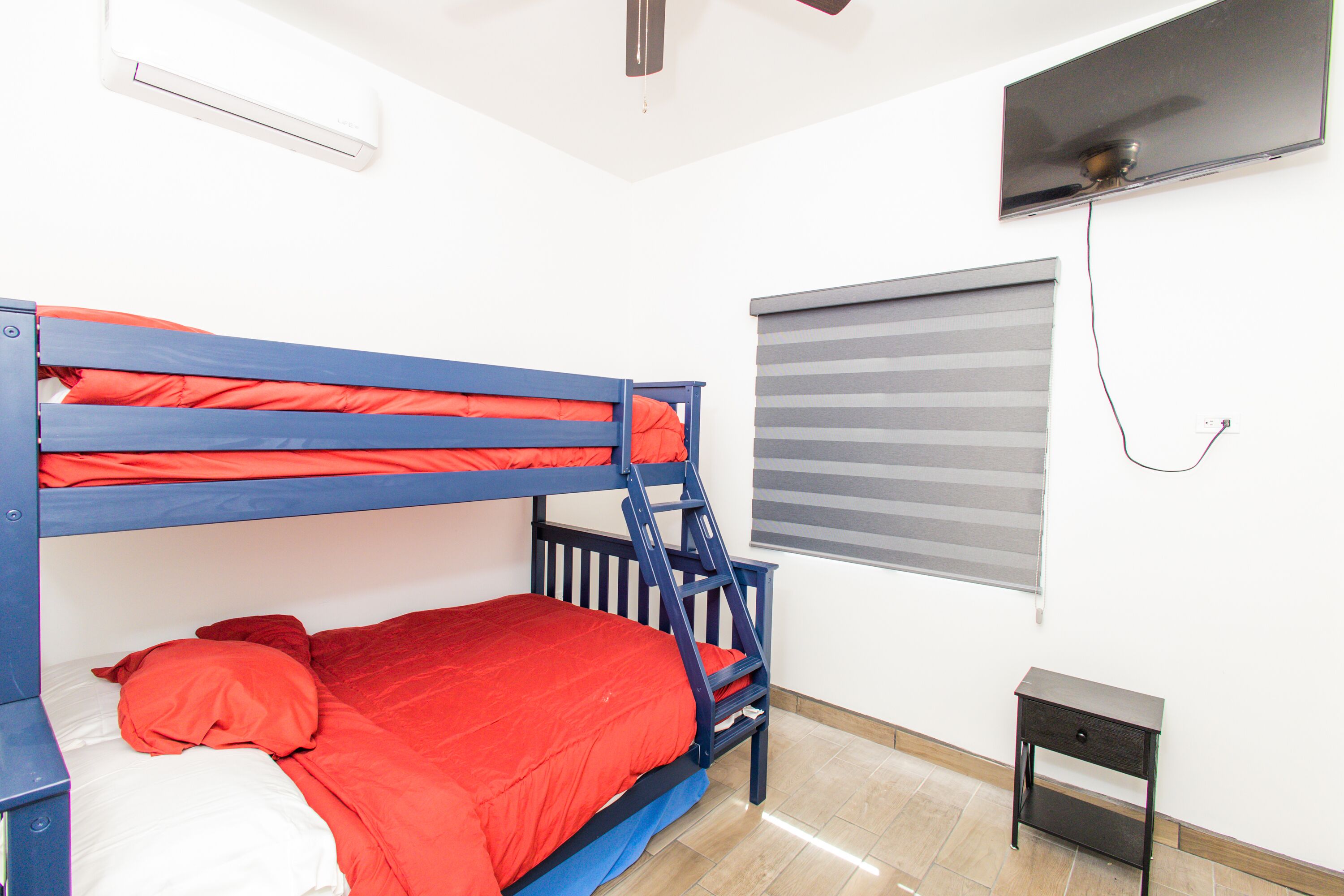 Een stapelbed met rode beddengoed, een klein nachtkastje, een aan de muur bevestigde televisie en een raam met zonwering.