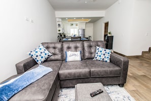 Uma sala de estar moderna com um sofá seccional cinza, duas almofadas decorativas estampadas, um controle remoto sobre uma mesa de centro e uma cozinha visível ao fundo.