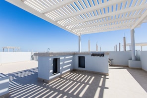 Um pátio ao ar livre com pérgola branca, área de churrasqueira integrada e vista para o mar.