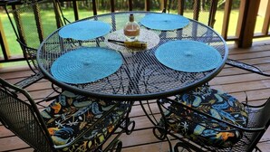 A round metal table with blue placemats and a bottle of water, surrounded by chairs with floral cushions, on a wooden deck with a view of trees. - Cottage at Lake Cumberland; 6m walk to water at Clearwater public boat ramp (Nancy)