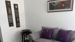 Una sala de estar con un sofá gris, cojines morados, una mesa de centro redonda con patas de metal, una mesita auxiliar y dos piezas de arte abstracto en la pared.