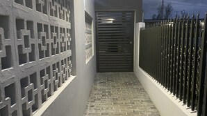 Un sendero pavimentado y estrecho conduce a una entrada con puerta, con un muro decorativo a un lado y una cerca metálica al otro.