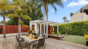 An outdoor patio with a wooden deck, a dining table with chairs, and a small white building in the background. - Newly Remodeled Charming and Contemporary Home (Santa Barbara)