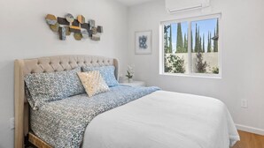 Une chambre à coucher comprenant un lit, un ventilateur de plafond, un appareil de climatisation, une fenêtre donnant sur la verdure et un mur orné de carreaux décoratifs.