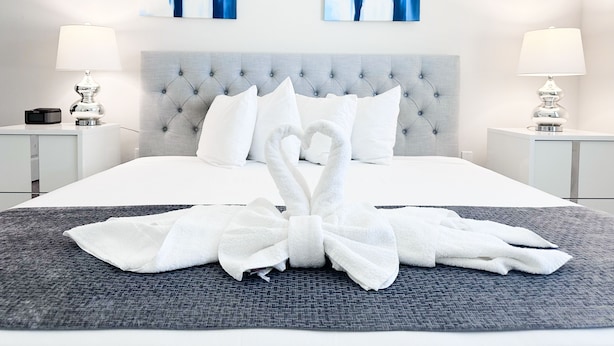 Ein modernes Hotelzimmer mit einem gepolsterten Kopfteil, einem ordentlich bezogenen Bett mit weißer Bettwäsche, zwei Nachttischen mit Lampen und abstrakter blauer Kunst an den Wänden.