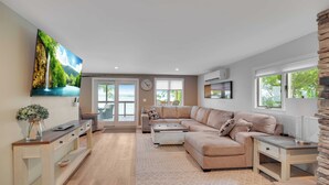 A modern living room with a large sectional sofa, a coffee table, and a TV mounted on the wall. There is a fireplace, a window with a view, and a door leading to an outdoor area.