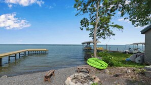 A lakeside setting with a wooden dock extending into the water, a picnic table, a fire pit, a kayak, and a covered area with seating.