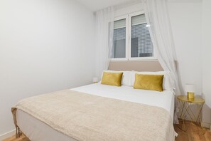 Ein Schlafzimmer mit Bett, einem Fenster mit Vorhängen, einem Nachttisch mit Lampe und einem kleinen runden Tisch ebenfalls mit Lampe.
