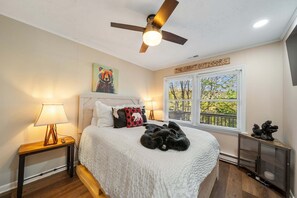 Une chambre à coucher avec un lit, un ventilateur de plafond, une table de chevet avec une lampe, une fenêtre avec des stores et un panneau accroché au mur.