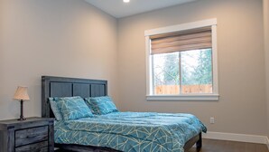 Un dormitorio con cama, mesita de noche, ventana con persianas y vista a árboles.