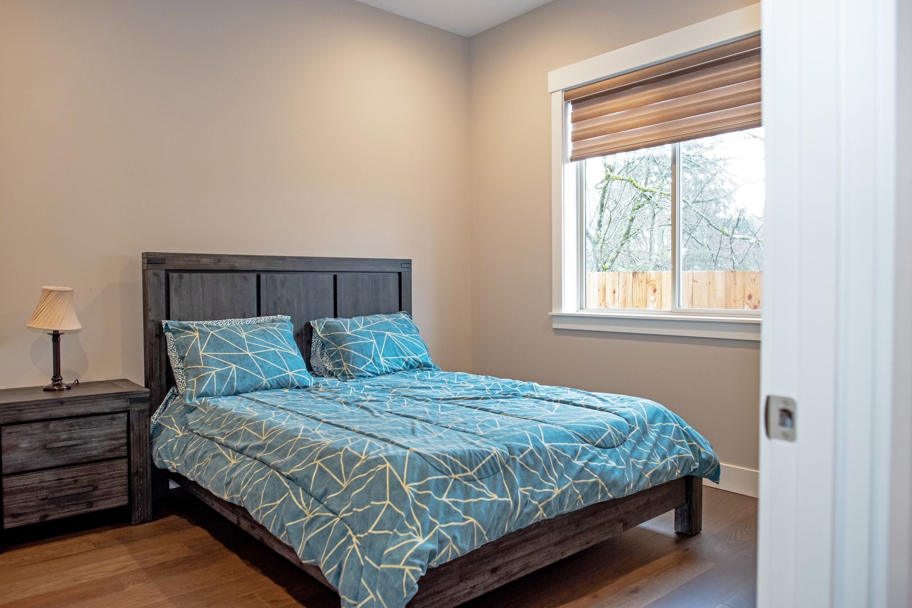 A bedroom with a bed, a nightstand, a window with blinds, and a view of trees outside.