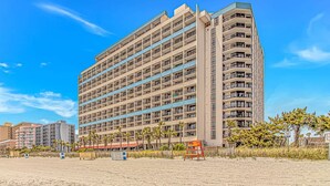 Een meerlaags hotel met balkons, nabij een strand met palmbomen.