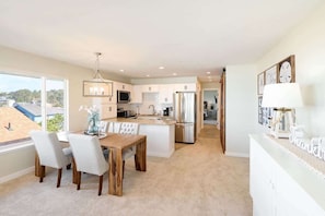 A modern kitchen with white cabinetry, a dining area with a wooden table and chairs, and a view of a residential area through large windows.