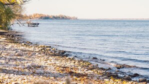 A rocky shoreline with a boat in the distance and a clear view of the water. - Historic Loyalist Lakehouse • Quiet Waterfront Escape • 3.5 Acres & 300’ Shore! (Prince Edward)