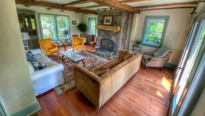 A spacious living room with wooden beams, a fireplace, and large windows. - Historic Loyalist Lakehouse • Quiet Waterfront Escape • 3.5 Acres & 300’ Shore! (Prince Edward)