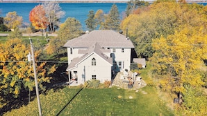 A white house with a grey roof, surrounded by trees and a body of water. - Historic Loyalist Lakehouse • Quiet Waterfront Escape • 3.5 Acres & 300’ Shore! (Prince Edward)
