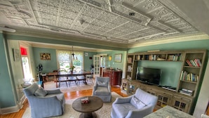 A spacious living room with a high ceiling, a dining area, and a library with bookshelves. - Historic Loyalist Lakehouse • Quiet Waterfront Escape • 3.5 Acres & 300’ Shore! (Prince Edward)