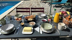 Ein Esstisch ist reichhaltig mit Frühstücksartikeln gedeckt, darunter Brot, Eier und Saft, in der Nähe eines Poolbereichs mit Liegestühlen und einem steinbelagten Weg.