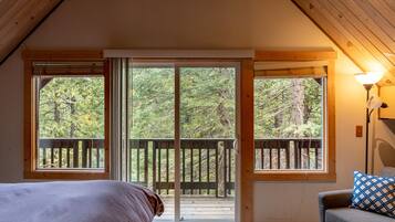 A bedroom with a large sliding glass door leading to a balcony with a wooden deck, a bed, a chair, and a lamp.