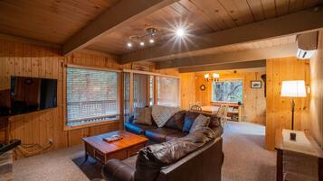 A cozy wooden cabin interior with a leather sofa, a dining table, and a television.