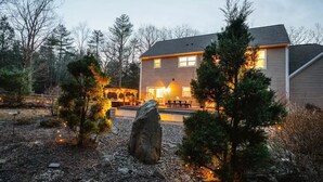 A two-story house with a patio area, surrounded by trees and a large rock.