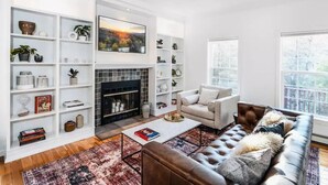 A living room with a fireplace, white shelves with decorative items, a brown leather sofa, and a large window.