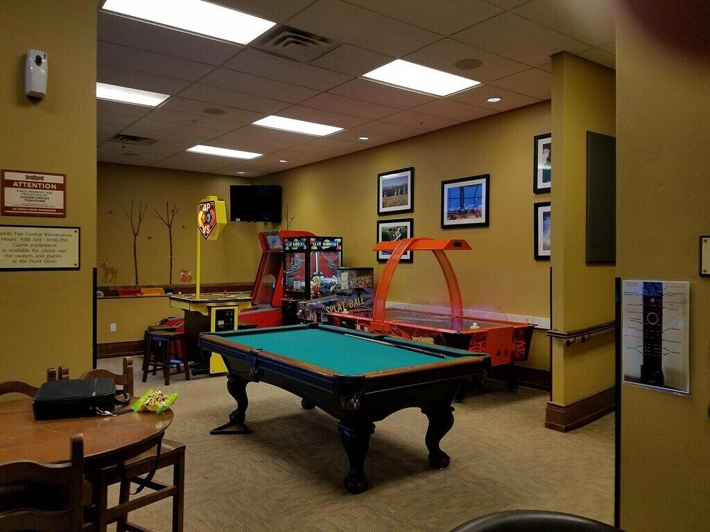 Une table de billard dans une pièce aux murs jaunes, avec un jeu de fléchettes et des jeux d’arcade.