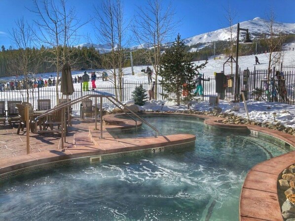 Un espace avec un bain à remous et une piscine, entouré d’une clôture, avec une montagne enneigée en arrière-plan.
