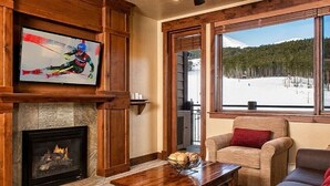 Un salón acogedor con chimenea, un televisor montado en la pared, un sofá cómodo y vistas a un paisaje nevado a través de amplios ventanales.