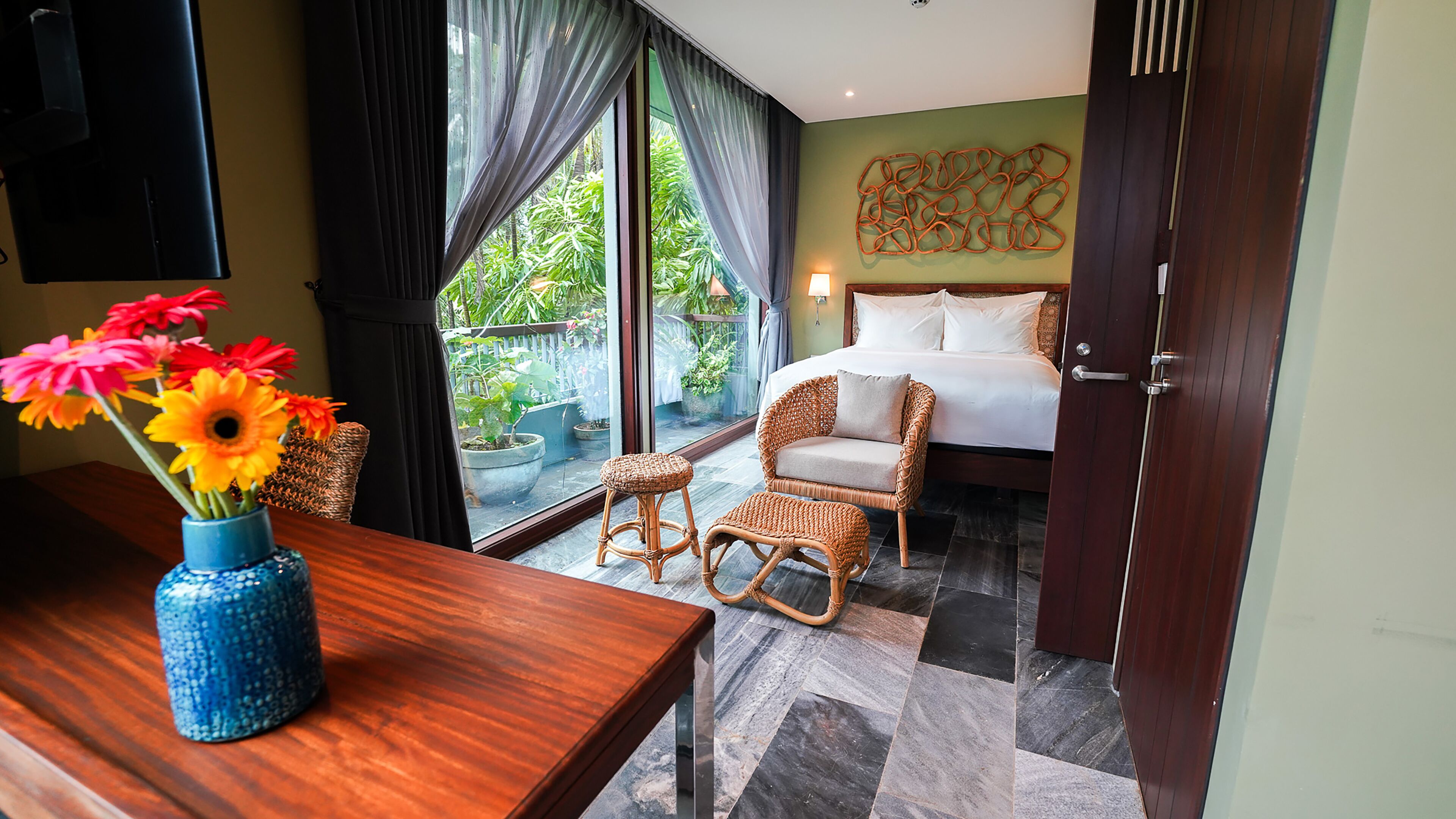 A hotel room with a bed, wicker furniture, a table with a vase of flowers, and a view of greenery outside.