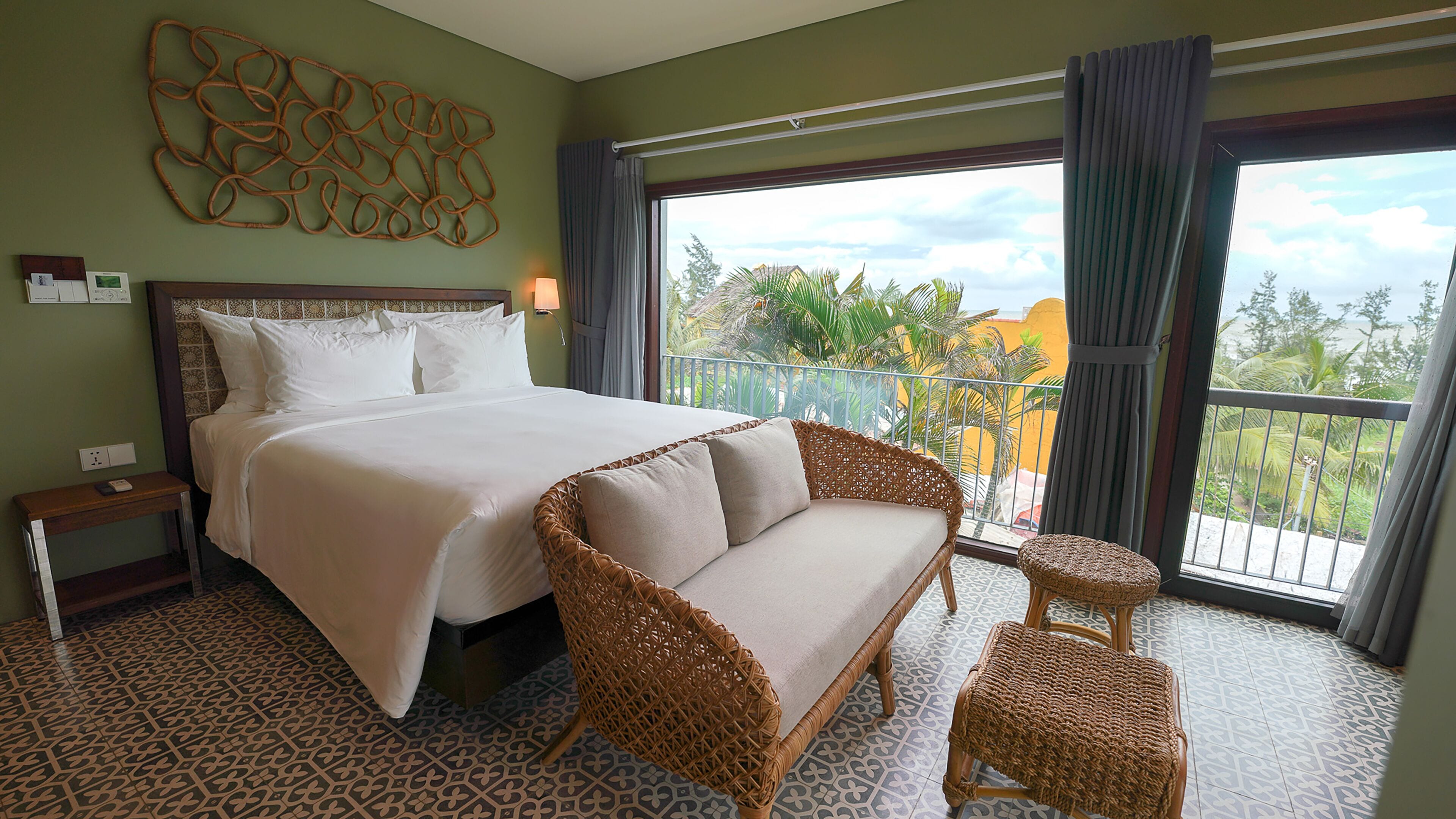 A hotel room with a large bed, a sofa, and a balcony with a view of palm trees and a beach.
