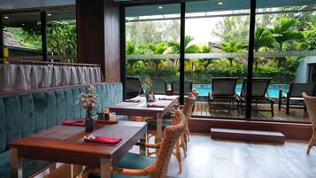 Un restaurante con vista a una piscina, mesas de madera, asientos acolchados y abundante vegetación.