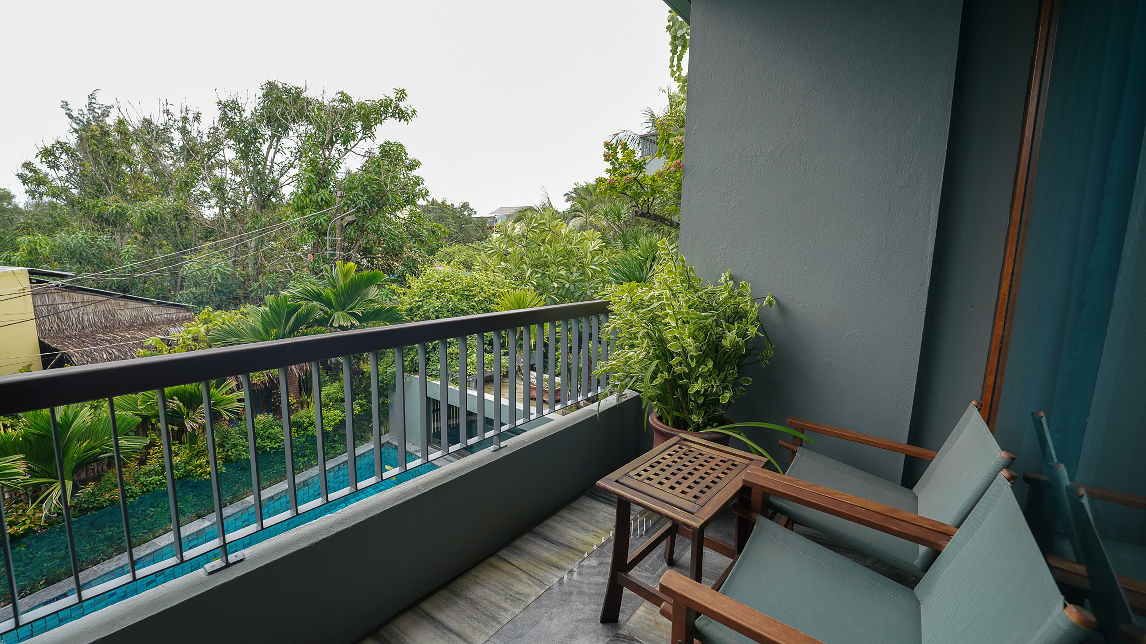 A balcony with a wooden chair and a small table, overlooking a swimming pool and lush greenery.