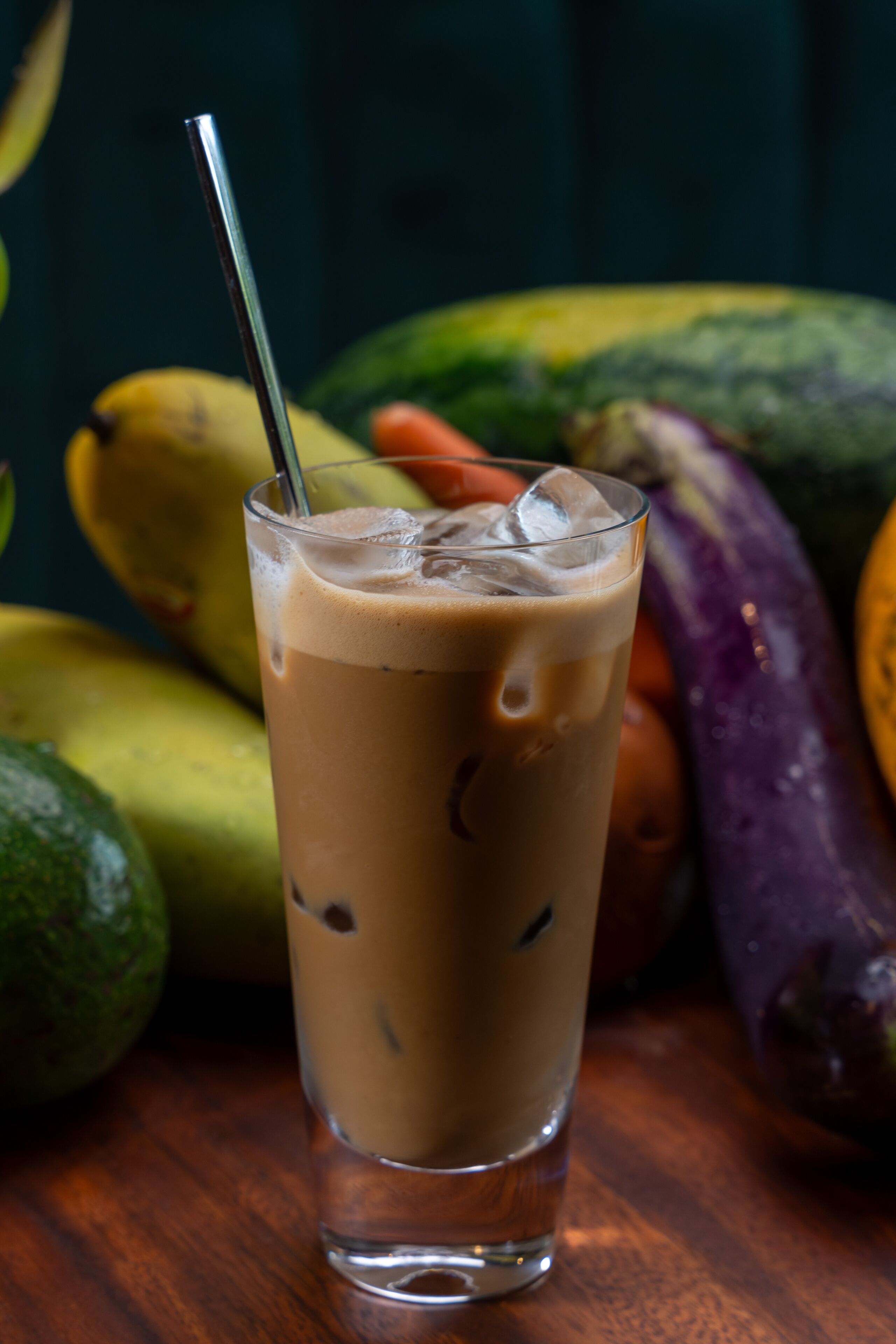 Un bicchiere alto di caffè ghiacciato con una cannuccia, circondato da diversi frutti come banane e avocado su un piano di legno.