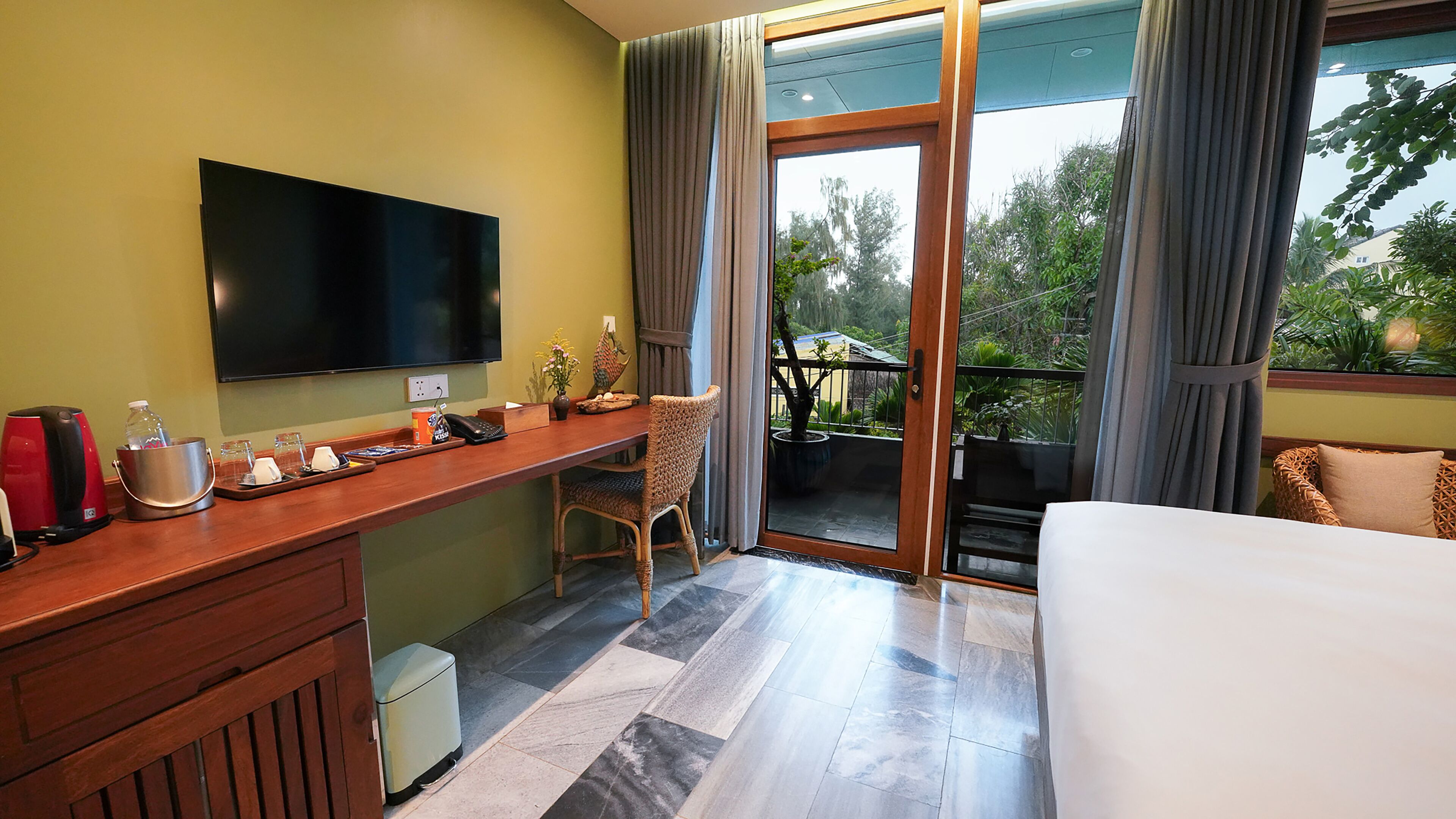 Habitación de hotel con escritorio de madera, televisión de pantalla plana, cama y balcón con vistas a la vegetación.