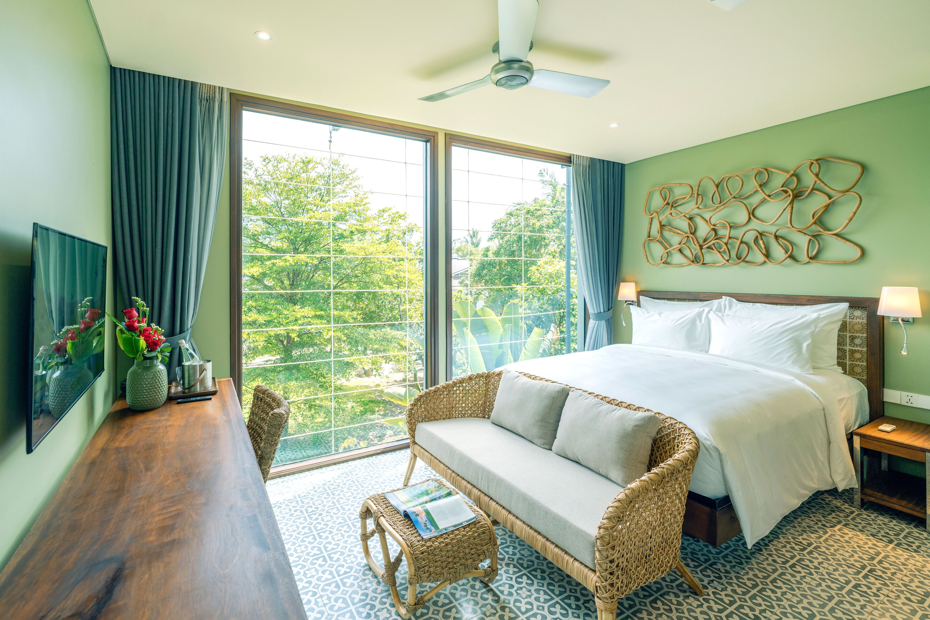 A hotel room with a large bed, a wicker sofa, a small table with a magazine, and a view of greenery through the windows.