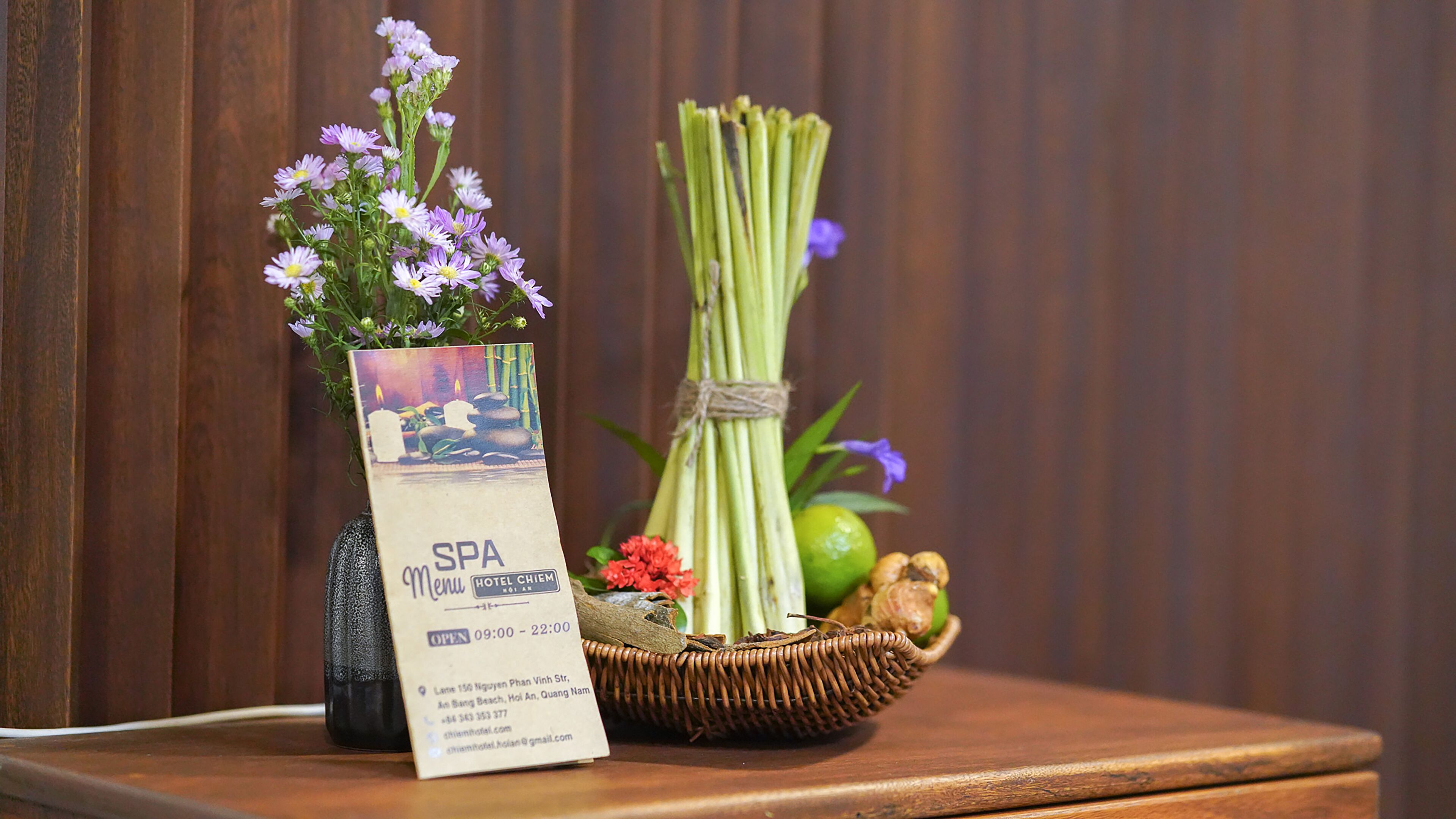 Un cartello del menù della spa, un cesto di vimini con cipollotti e un fiore rosso, e un vaso con fiori viola su un tavolo di legno.