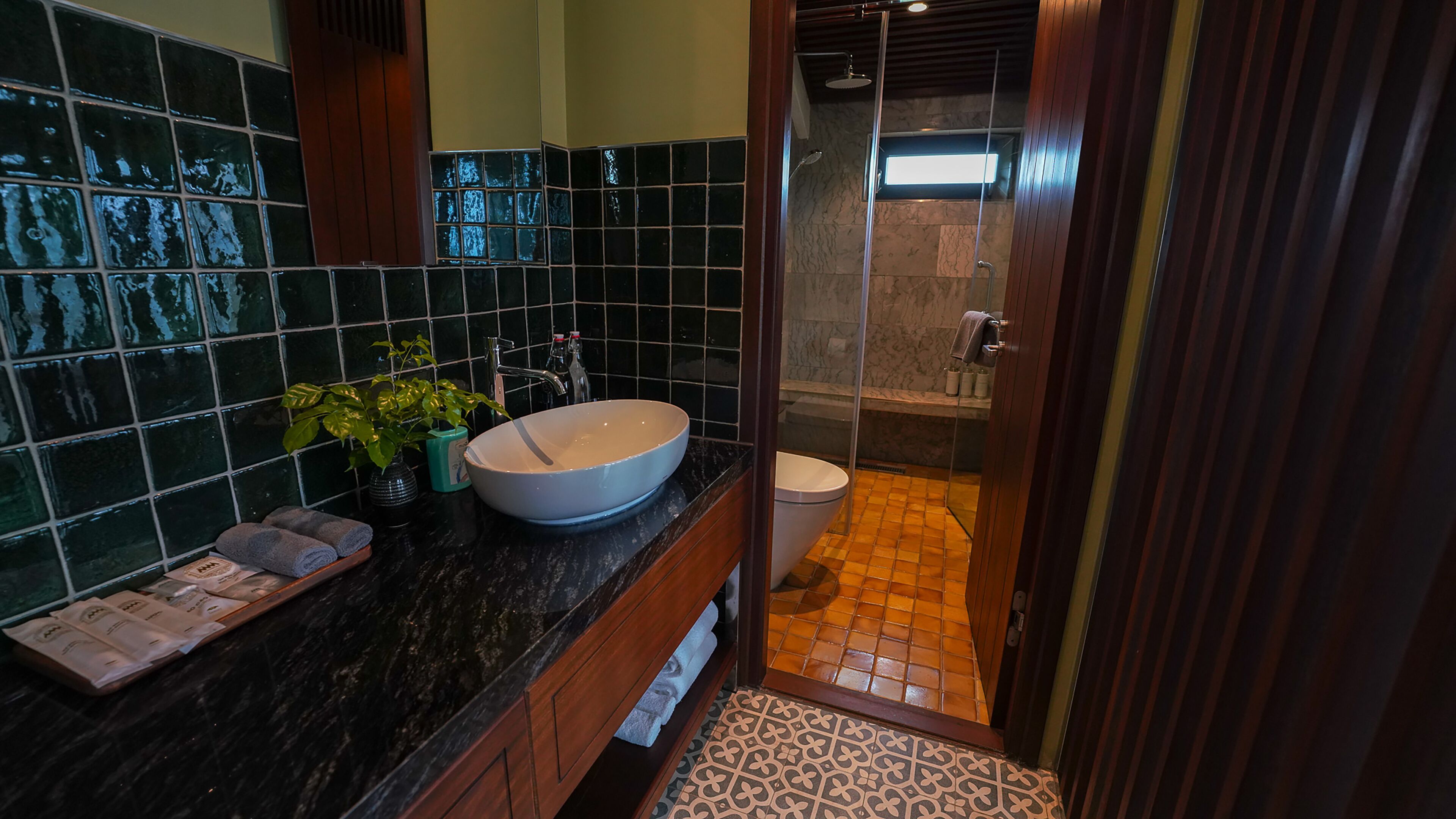 A bathroom with a large white sink, dark tiled walls, a wooden vanity, and a shower area with orange tiles.