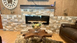 Un salon chaleureux avec une cheminée en pierre, une table basse en bois sur laquelle est posé un panier de fruits, et un canapé en cuir marron.