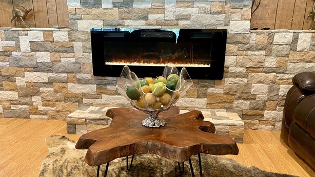 Une cheminée en pierre avec un feu de bois, une table en bois sur laquelle est posé un panier de fruits, et un canapé en cuir.