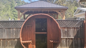 Une structure en forme de tonneau en bois, dotée d’une porte, entourée d’une clôture en bois et comprenant un espace abrité sous un toit.